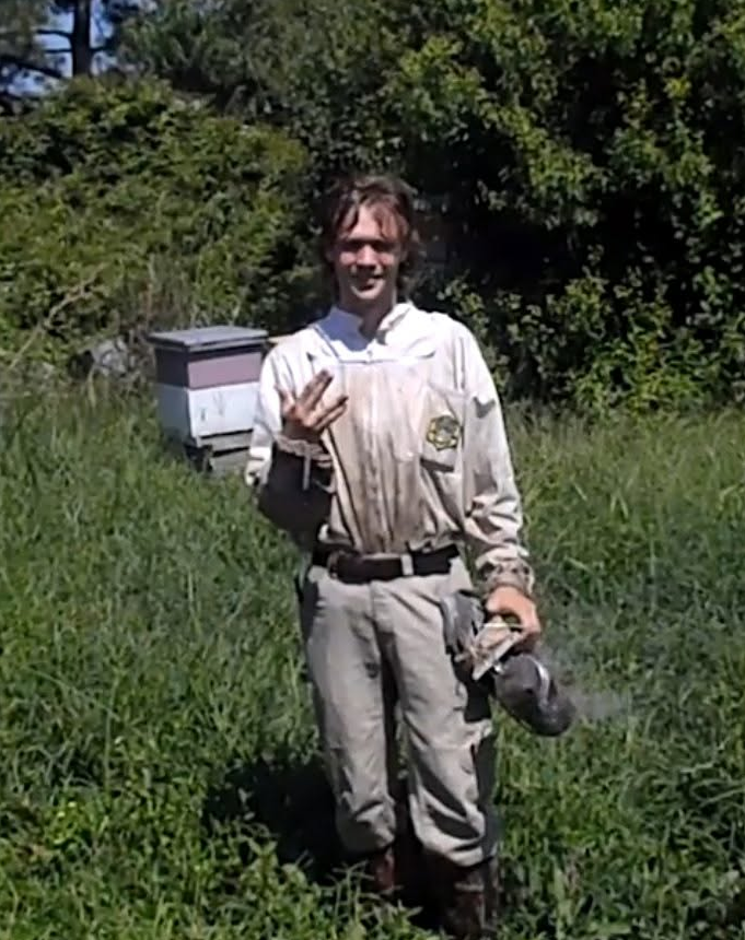 Chase Cruz in a beekeeping suit, standing in tall grass beside a hive